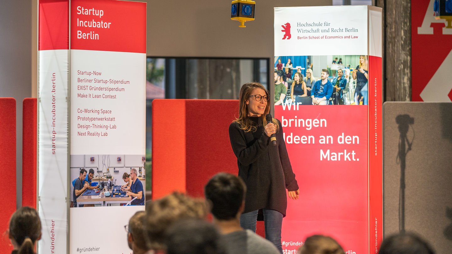 Mitarbeitende des Startup Incubator Berlin stellt auf einer Messebühne mit dem Mikro in der Hand das Berliner Startup Stipendium vor. Foto: Startup Incubator Berlin, Matthias Grytzka