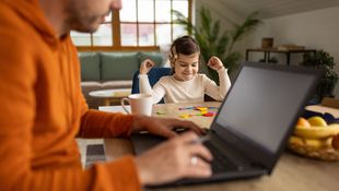 Mann arbeitet am Laptop, im Hintergrund sitzt ein Kind und malt