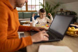 Mann arbeitet am Laptop, im Hintergrund sitzt ein Kind und malt