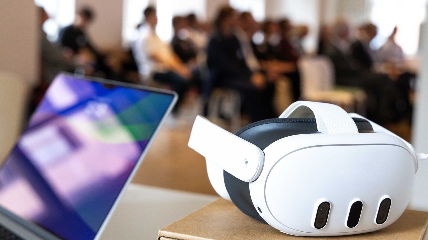 Laptop und VR Brille liegen auf einem Tisch, Menschen im Hintergrund