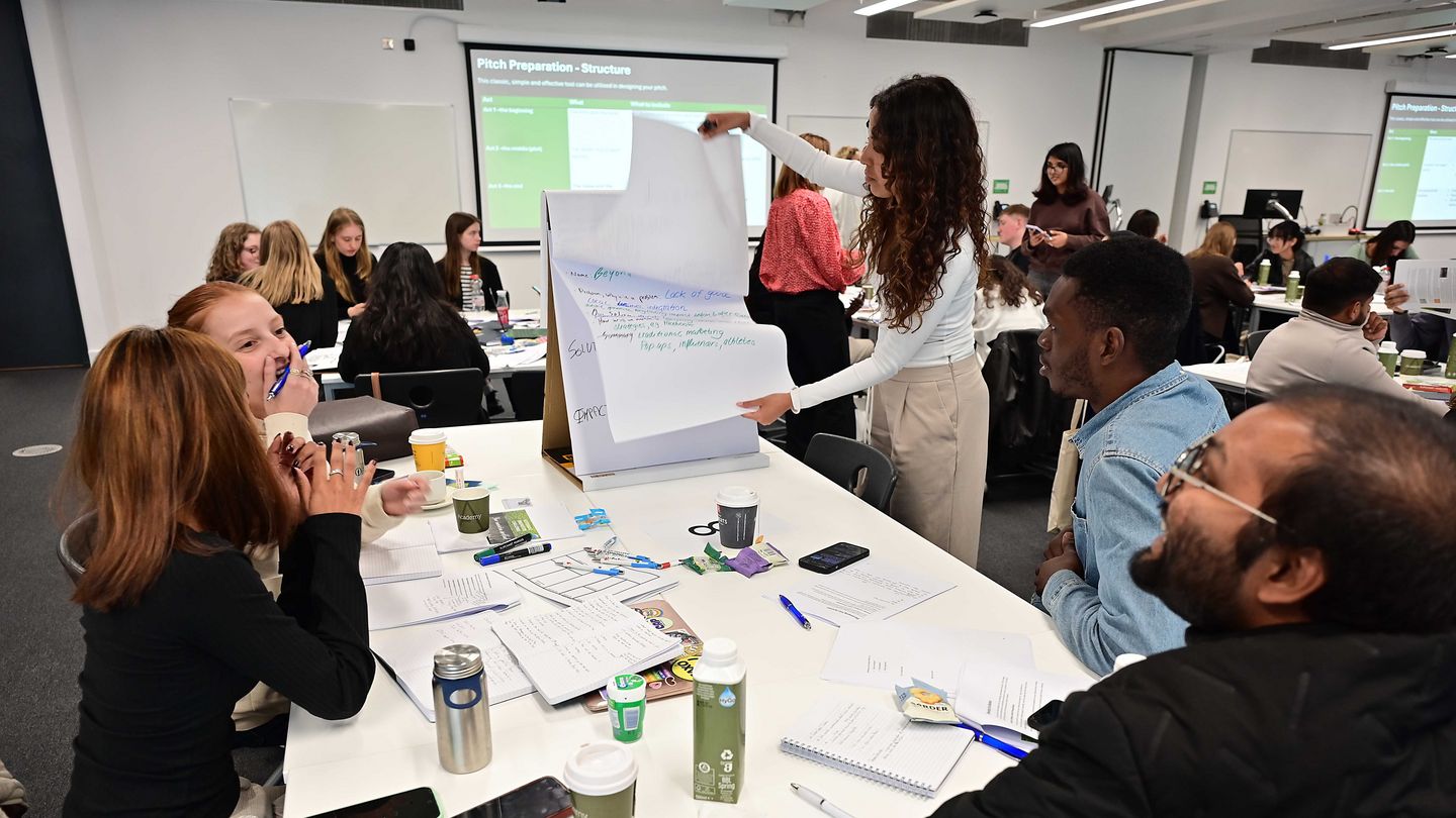 Studierende an Arbeitstischen und einem Flipchart