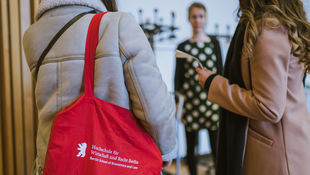 [Translate to English:] Zwei Studentinnen mit einer roten HWR-Tasche im Gespräch mit einer Studienberaterin des Fachbereichs Allgemeine Verwaltung. Foto: Oana Popa-Costea