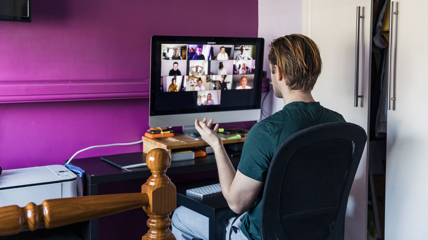 A view over the shoulder: A student sits in front of his PC at home and looks at a video conference on the screen, which is placed in front of a purple wall. Photo: © SolStock/ E+/ Getty Images