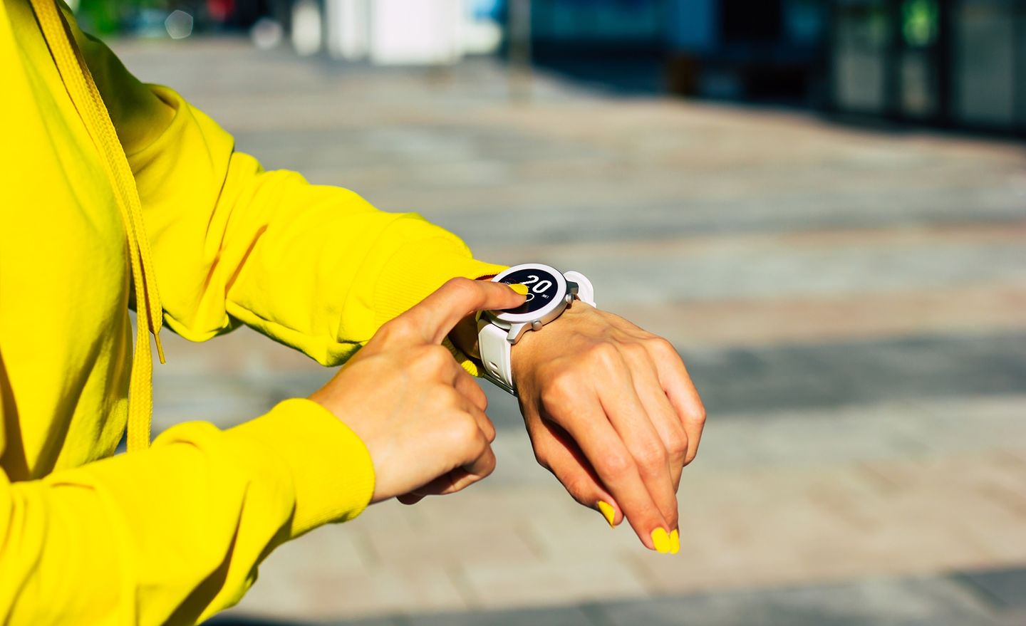 Eine Studierende im gelben Sweatshirt deutet mit dem rechten Zeigefinger auf ihre Smartwatch am linken Handgelenk. Die Smartwatch zeigt ein Datum an. Foto: © Povozniuk/Getty Images/iStockphoto