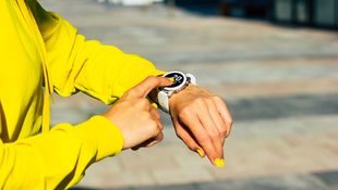 Eine Studierende im gelben Sweatshirt deutet mit dem rechten Zeigefinger auf ihre Smartwatch am linken Handgelenk. Die Smartwatch zeigt ein Datum an. Foto: © Povozniuk/Getty Images/iStockphoto