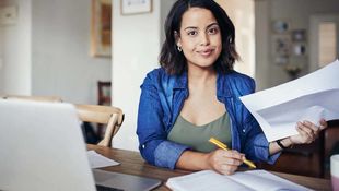 Eine junge Frau mit dunklen Haaren studiert am Esstisch vor dem geöffneten Laptop und macht sich Notizen. Foto:  iStock | mapodile