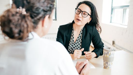 Studienberatung: Beraterin mit Brille und lockigem Haar am Schreibtisch im Gespräch mit einer Studierenden mit Pferdeschwanz. Foto: © kate_sept2004/E+/Getty Images