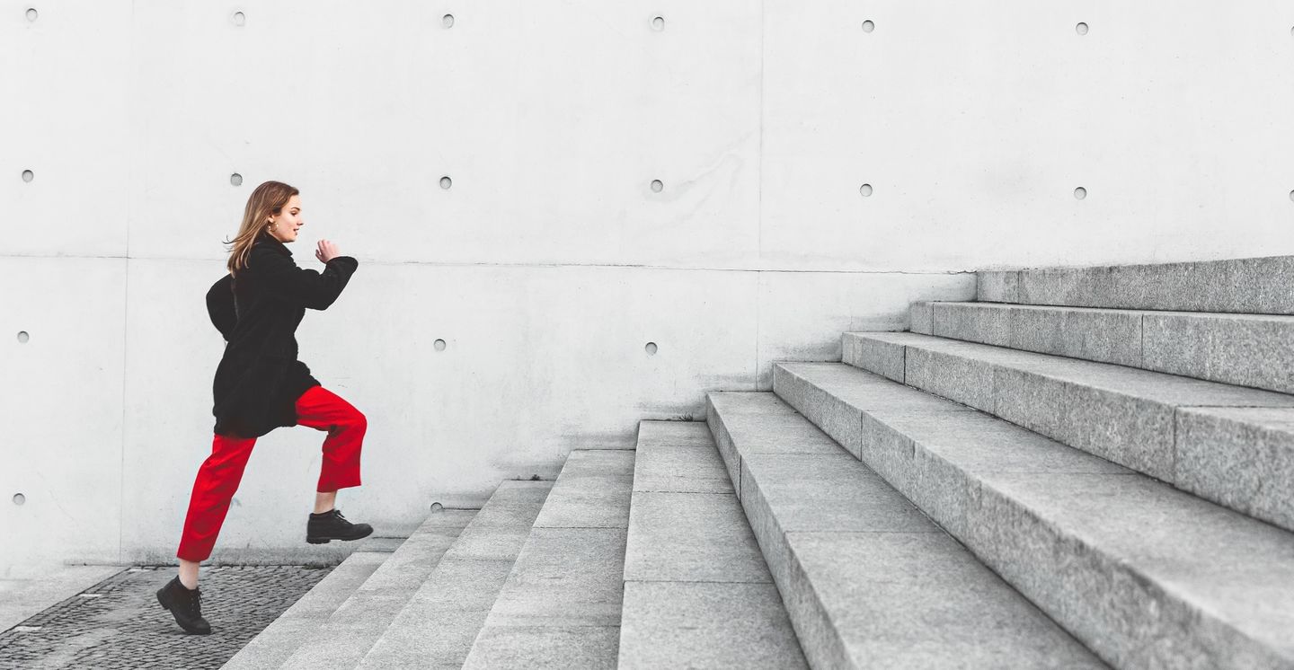 Dein Studium an der HWR Berlin: Eine Studierende in schwarzer Jacke und roten Hosen eilt eine Treppe aus Sichtbeton hinauf. Foto: © Nikada/E+/Getty Images