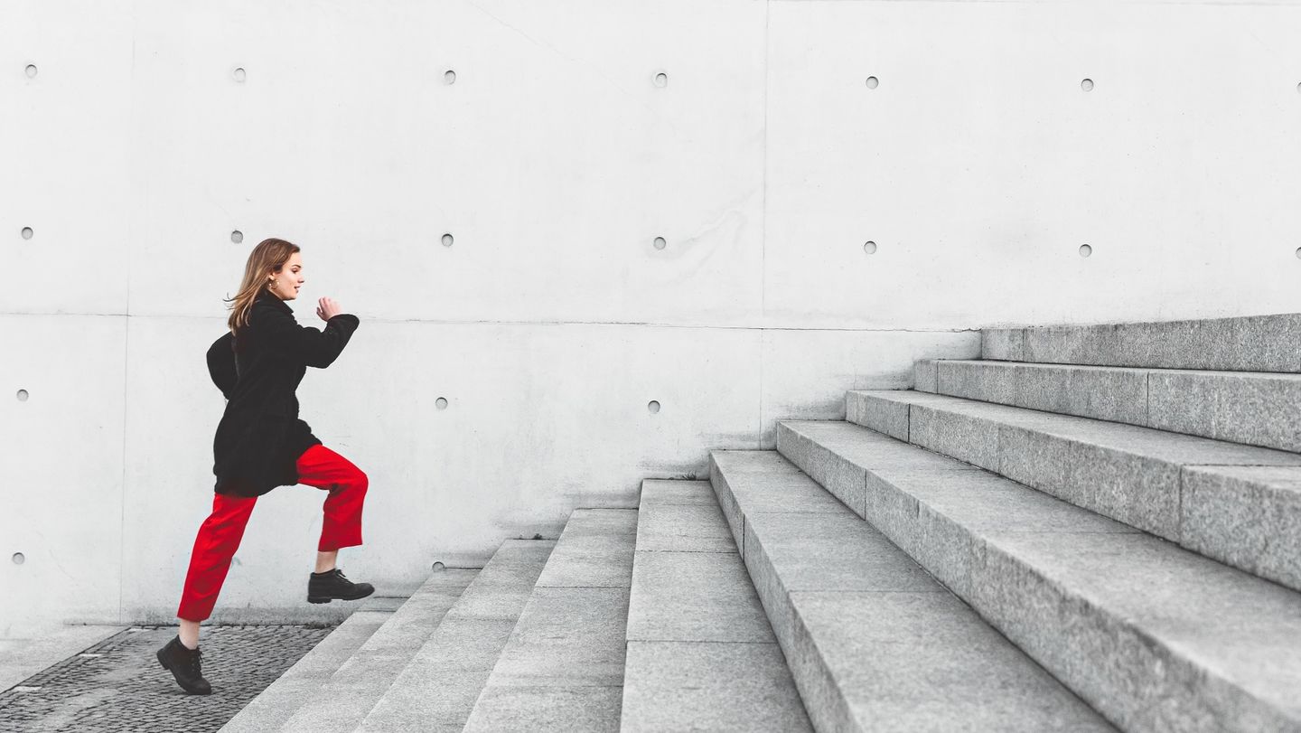 Dein Studium an der HWR Berlin: Eine Studierende in schwarzer Jacke und roten Hosen eilt eine Treppe aus Sichtbeton hinauf. Foto: © Nikada/E+/Getty Images