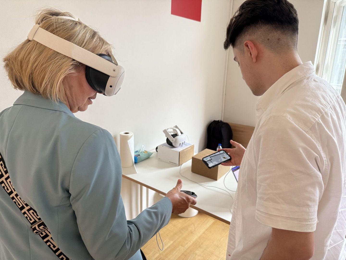 Eine Frau trägt eine Virtual-Reality-Brille und hält einen Controller in der Hand. Neben ihr steht ein junger Mann, der auf sein Smartphone schaut und ihr die Anwendung erklärt. Beide befinden sich in einem hellen Raum mit einem Tisch, auf dem weiteres technisches Equipment liegt.
