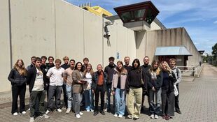 Gruppe von jungen Erwachsenen und zwei älteren Personen steht vor einer hohen Betonmauer mit einem Wachturm und Überwachungskamera unter blauem Himmel