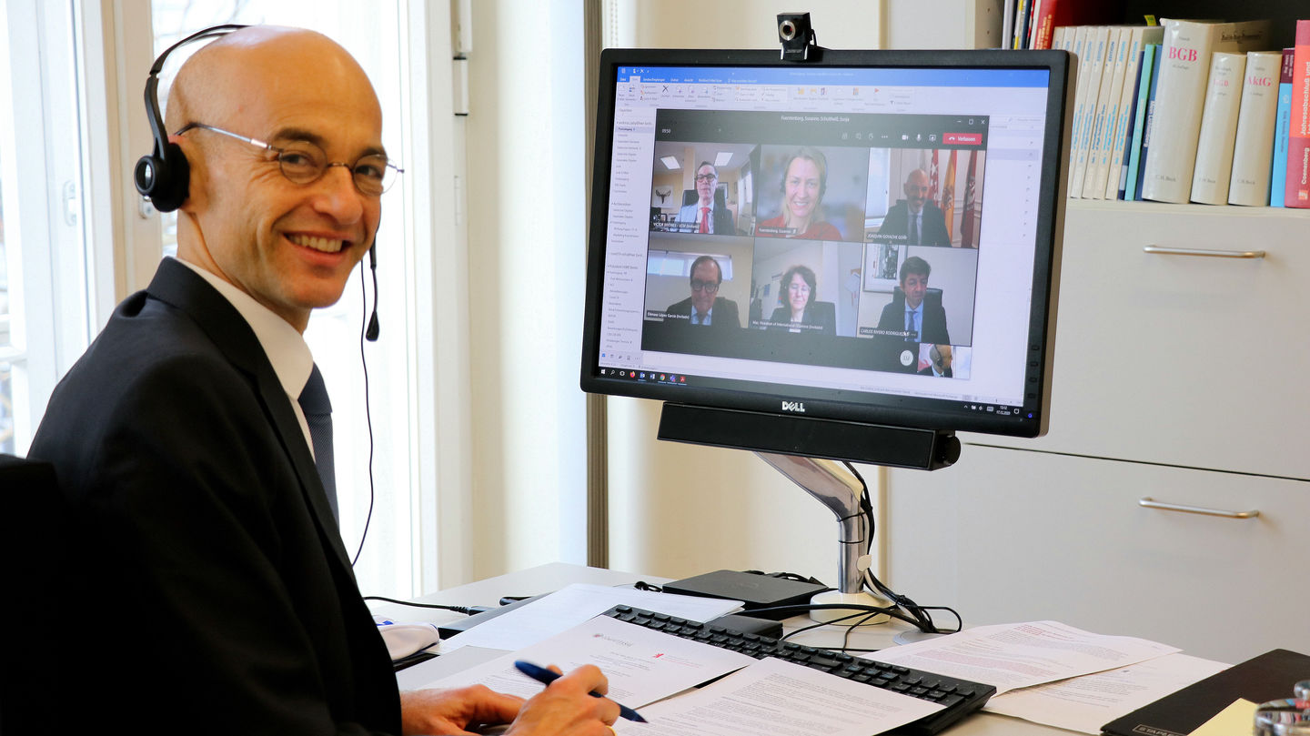 Mann sitzt am Schreibtisch vor dem Bildschirm, auf dem eine Videokonferenz läuft.