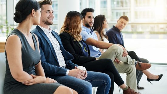 Stellenangebote: Das Foto zeigt drei weibliche und drei männliche Jobinteressierte unterschiedlicher Ethnien, die auf Stühlen in einer Reihe sitzen und warten. Foto: © Mikolette/E+/Getty Images