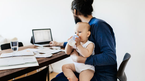 A young father holds an infant with a baby bottle on his lap and at the same time works on his laptop. Photo: © TommL/GettyImages/E+