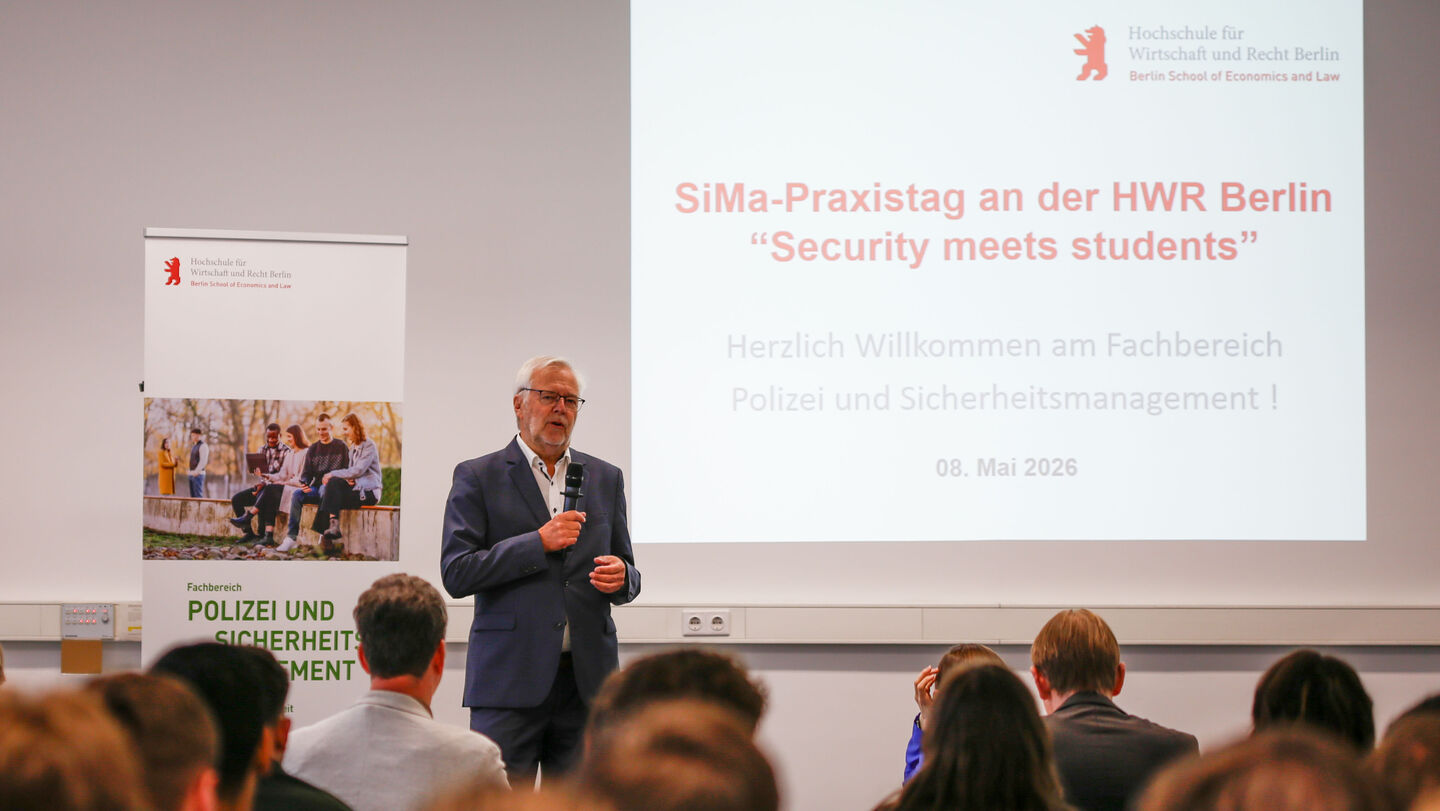 Vortragender steht vor Leinwand mit Präsentation zum Thema "SiMa-Praxistag an der HWR Berlin Security meets students" vor Publikum in einem Seminarraum.