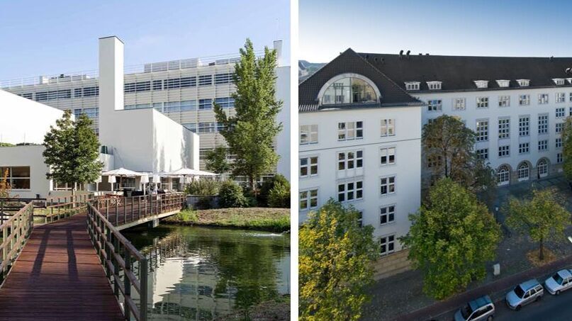 Links ein moderner Gebäudekomplex mit Holzsteg über Wasser, rechts ein historisches mehrstöckiges Gebäude mit Satteldach und parkenden Autos davor.