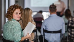 Fachbereichsrat diskutiert bei einer Sitzung. Eine Studierende mit roten Locken hält die Beratungsunterlagen in der Hand und dreht sich lächelnd zur Kamera. Foto: © skynesher/Getty Images/E+