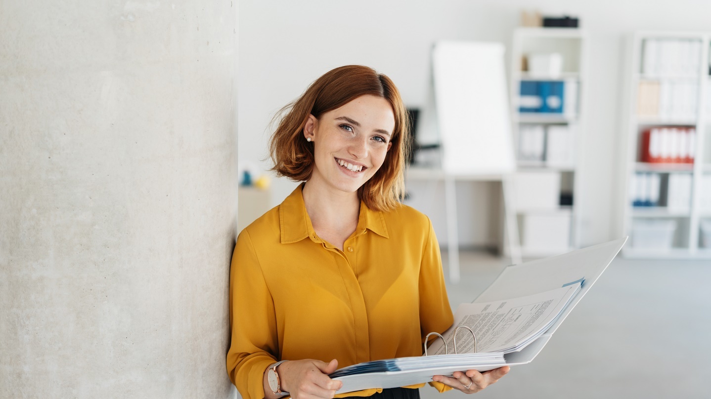 Junge Frau mit gelber Bluse vor grauer Wand mit einem Ordner in der Hand. Foto: © stockfour/Getty Images/iStockphoto