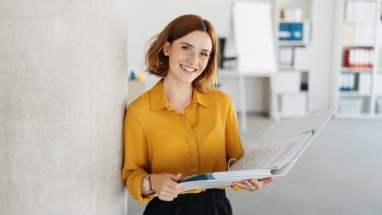 Junge Frau mit gelber Bluse vor grauer Wand mit einem Ordner in der Hand. Foto: © stockfour/Getty Images/iStockphoto