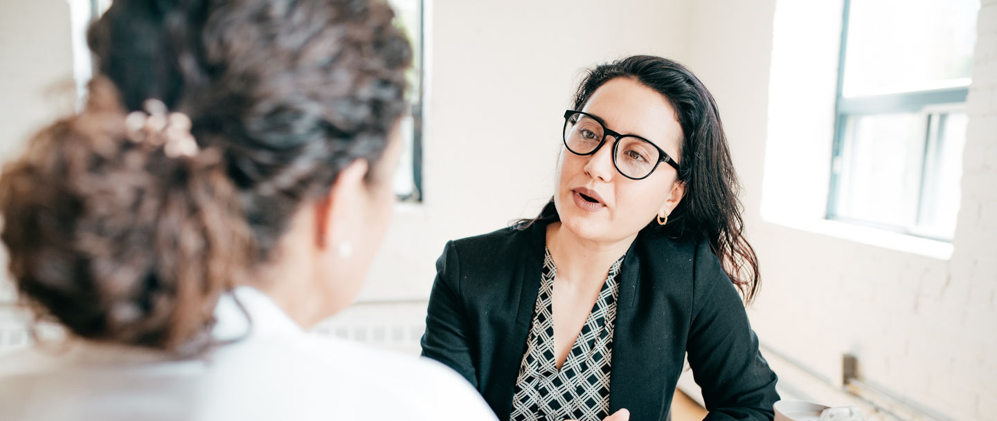 Studienberatung: Beraterin mit Brille und lockigem Haar am Schreibtisch im Gespräch mit einer Studierenden mit Pferdeschwanz. Foto: © kate_sept2004/E+/Getty Images