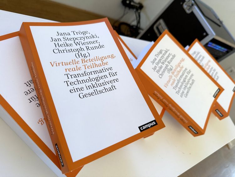 Mehrere Exemplare des Sammelbands „Virtuelle Beteiligung, reale Teilhabe. Transformative Technologien für eine inklusive Gesellschaft“ liegen auf einem weißen Tisch. Die Bücher haben einen weißen Umschlag mit orangefarbenem Rand und schwarzem sowie orangefarbenem Text.