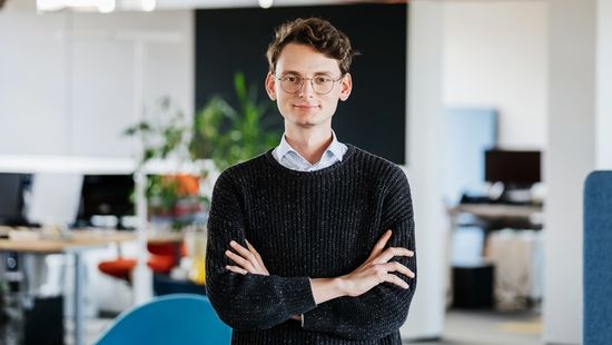 Junger Mann mit Brille steht mit überkreuzten Armen vor einem Bürohintergrund.
