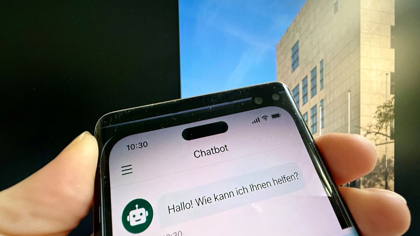 Hand hält Smartphone mit Chatbot-App, auf dem Bildschirm steht 'Hallo! Wie kann ich Ihnen helfen?'