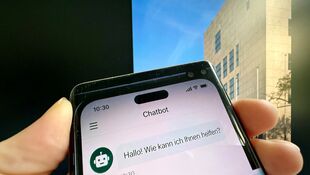 Hand hält Smartphone mit Chatbot-App, auf dem Bildschirm steht 'Hallo! Wie kann ich Ihnen helfen?'