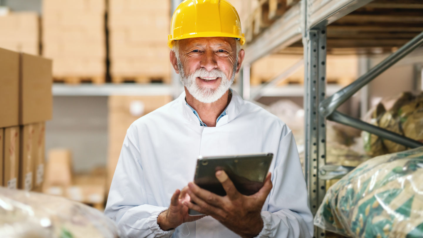 Forschung an der HWR Berlin: Ein älterer Industriearbeiter im Overall und  mit gelbem Helm bewegt sich zwischen Lagerregalen und hält ein Tablet. Foto: © dusanpetkovic/Getty Images/iStockphoto