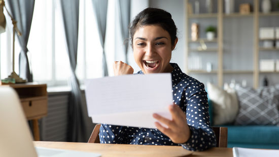 Studierende mit dunklen Haaren sitzt am Schreibtisch und freut sich über eine schriftliche Zusage für ein Stipendium. Foto: © fizkes/Getty Images/iStockphoto