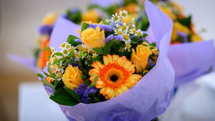 Blumenstrauß mit gelben Rosen, orange-gelber Gerbera, kleinen weißen Blüten und lila Blüten, eingewickelt in hellviolettes Papier