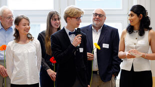 Gruppe von Menschen in formeller Kleidung, eine Person hält ein Mikrofon und eine gelbe Blume