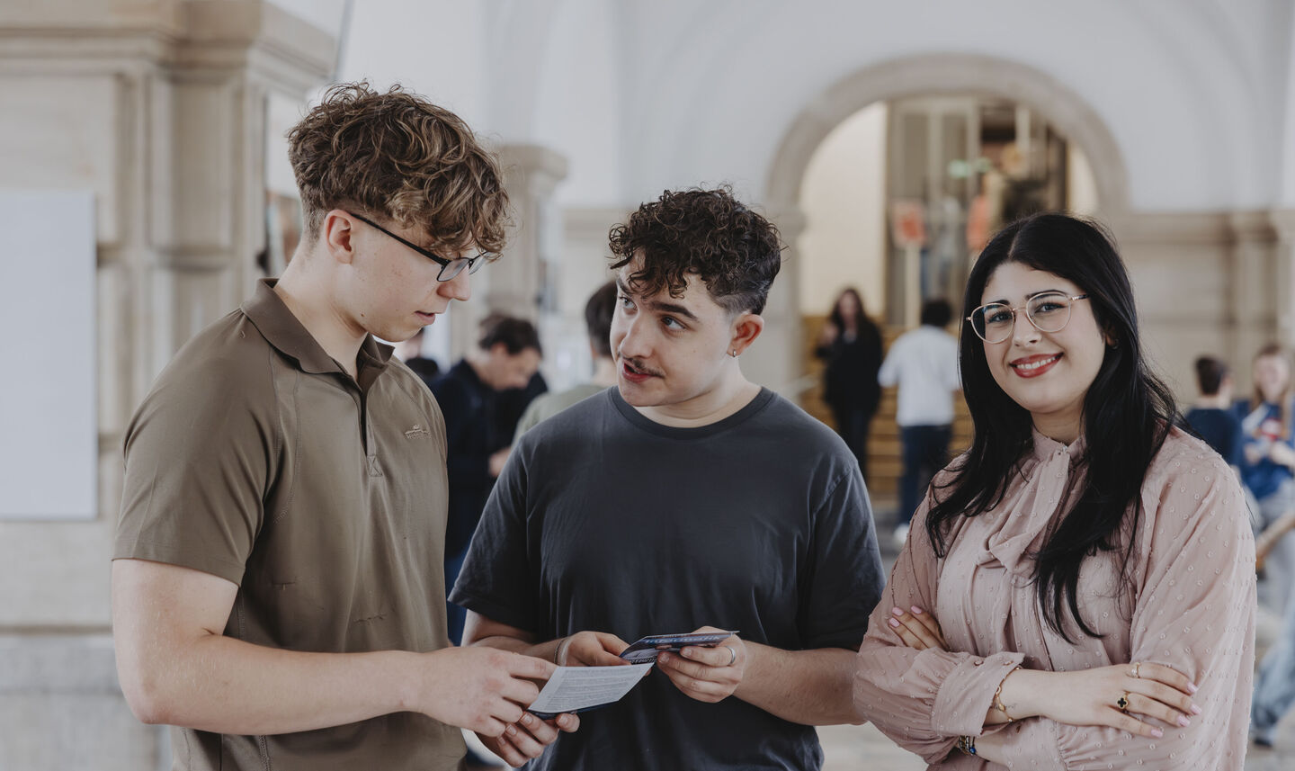 Drei junge Menschen stehen in einem hellen Innenraum, zwei halten Papiere, eine Person hat die Arme vor der Brust verschränkt
