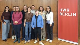 Gruppe von neun Personen steht vor einer Holzpaneelwand mit einer Uhr und einem roten Banner mit der Aufschrift 'HWR BERLIN'