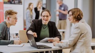 Drei Personen sitzen gemeinsam an einem Tisch um einen Laptop. 