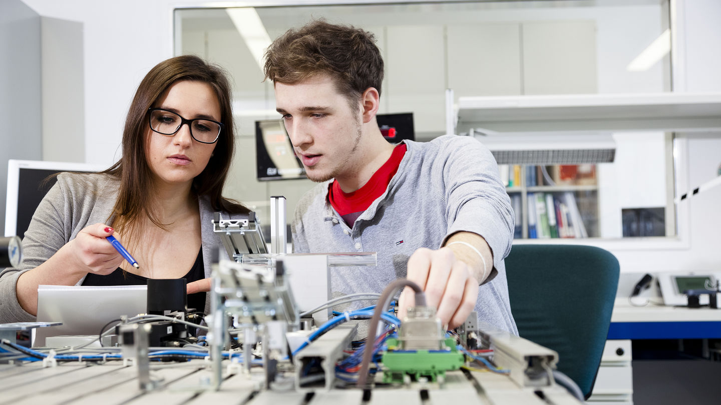Zwei Studierende sitzen in einem technischen Labor an einem Experiment.