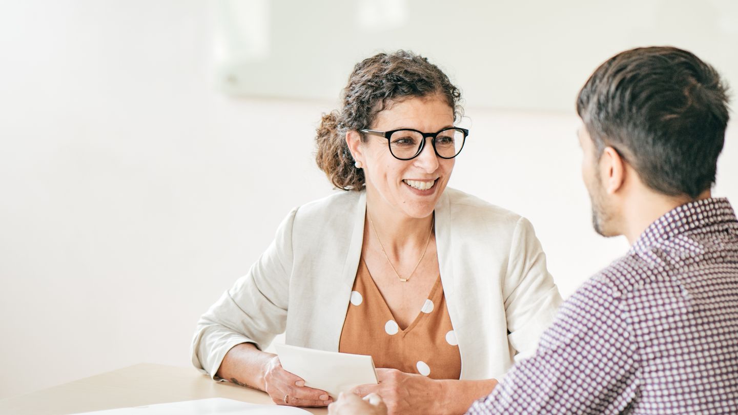 Student counselling: Counsellor with curly hair and glasses at the table talking to a student in a plaid shirt. Photo: © kate_sept2004/E+/Getty Images