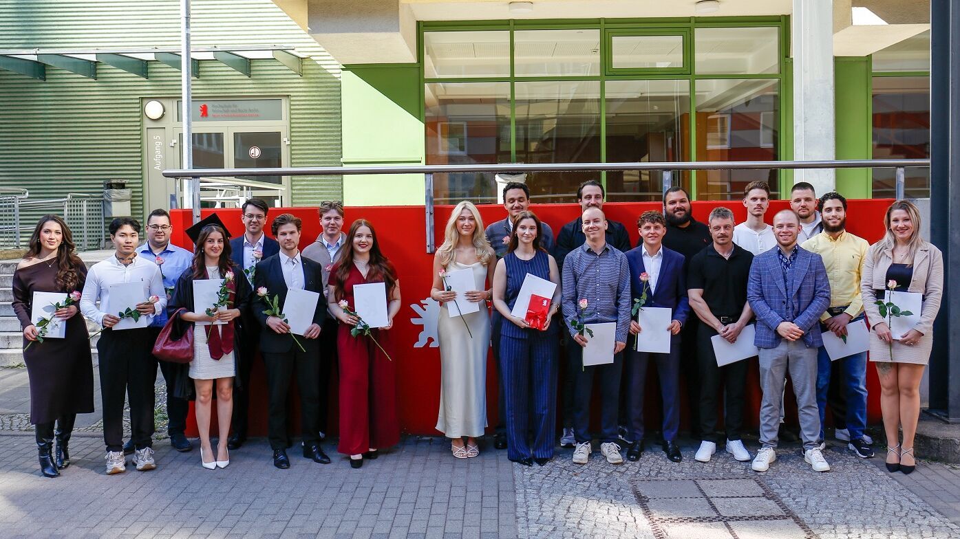 Gruppe von jungen Erwachsenen steht vor einem Gebäude, viele halten Urkunden und einzelne Rosen in der Hand.