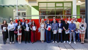 Gruppe von jungen Erwachsenen steht vor einem Gebäude, viele halten Urkunden und einzelne Rosen in der Hand.