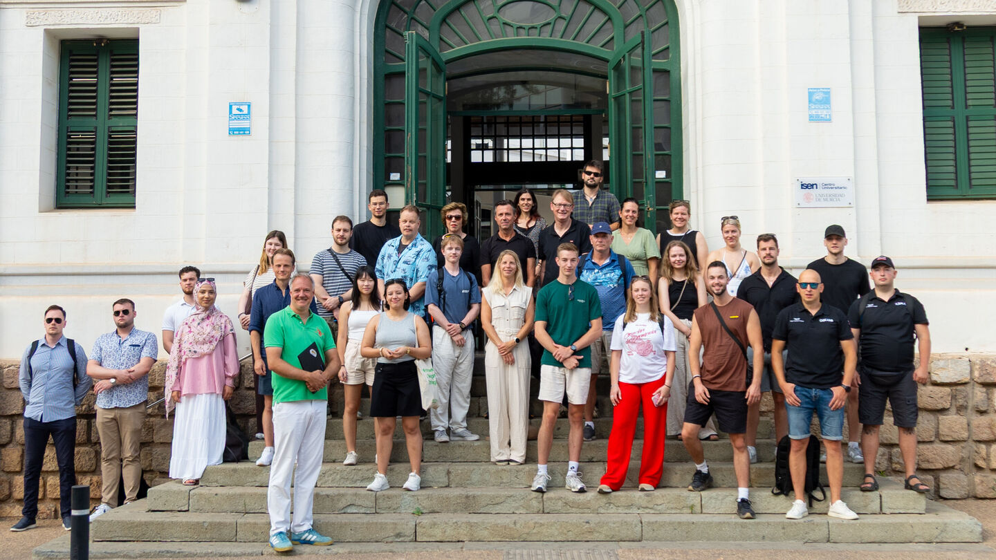 Gruppe von Menschen unterschiedlichen Alters und Geschlechts steht auf einer Treppe vor einem großen Gebäude mit grünem Tor und weißen Wänden