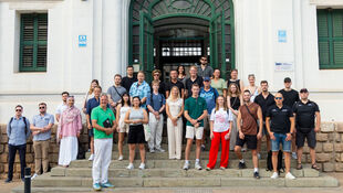 Gruppe von Menschen unterschiedlichen Alters und Geschlechts steht auf einer Treppe vor einem großen Gebäude mit grünem Tor und weißen Wänden