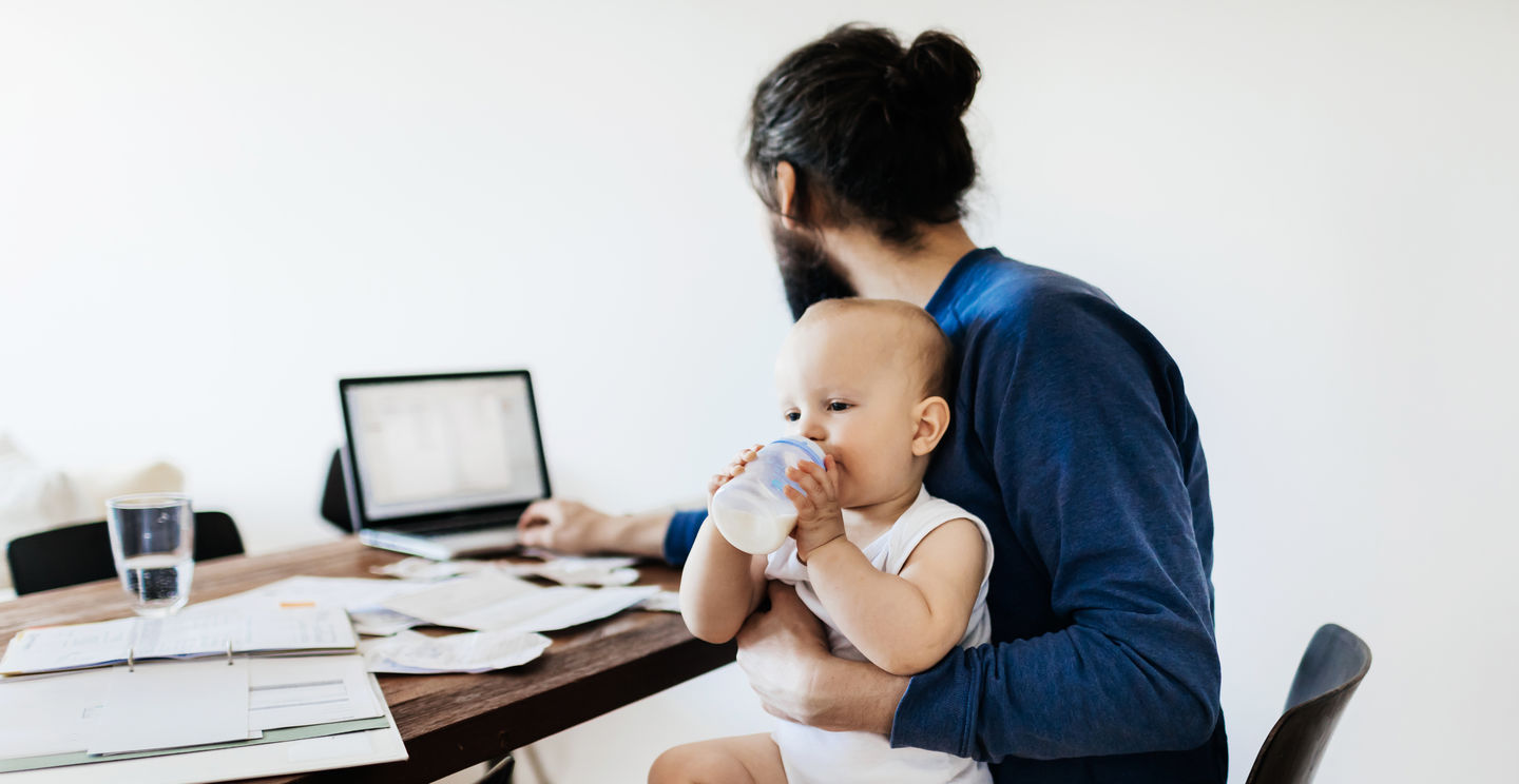 Studium mit Kind oder Familie: Junger Vater studiert zu Hause am Schreibtisch mit seinem Kleinkind auf dem Schoß, das aus einer Milchflasche trinkt. Foto: © TommL/GettyImages/E+