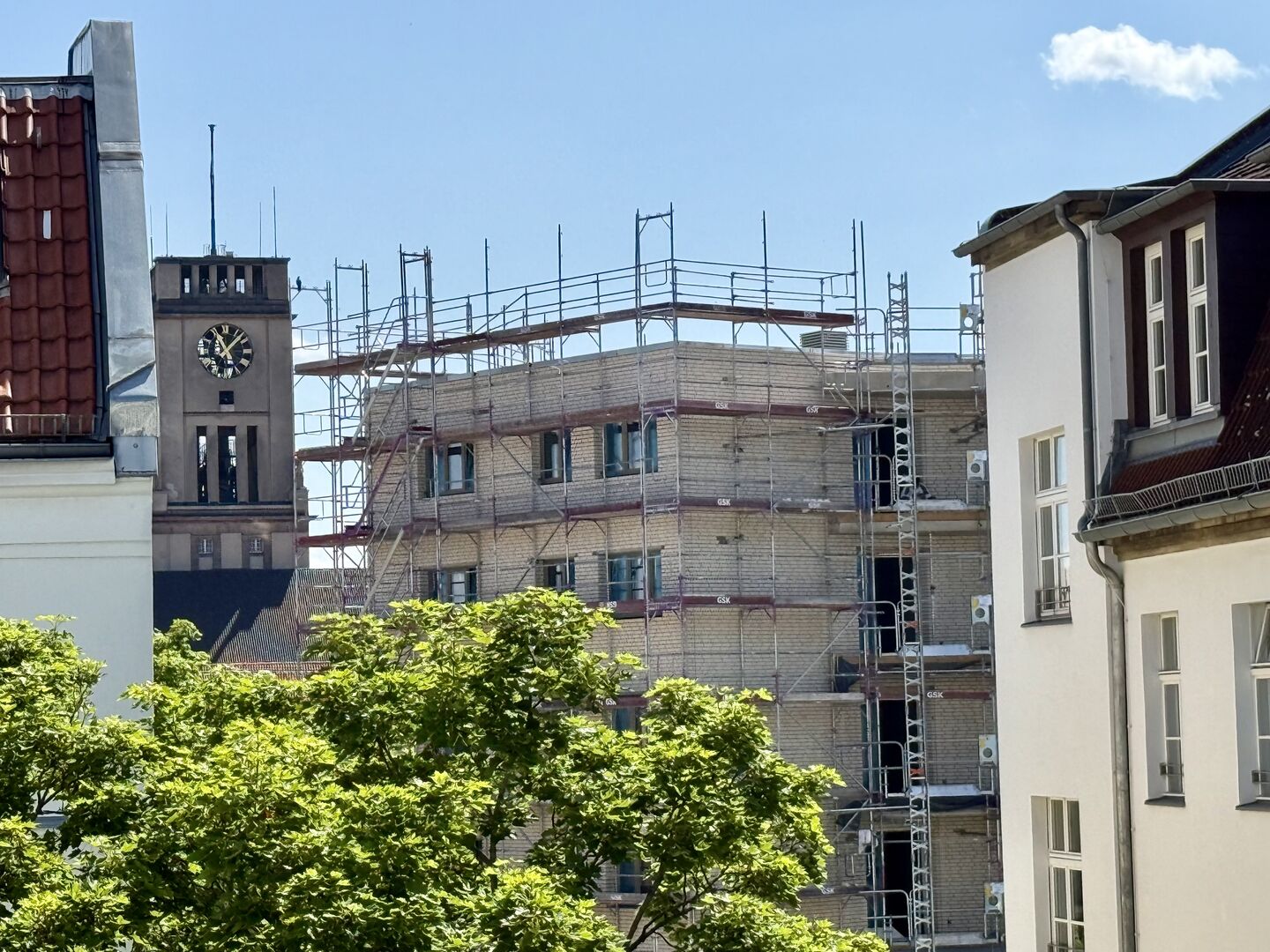 Blick auf ein mit Gerüst umgebenes Gebäude zwischen anderen Häusern und einem Kirchturm mit Uhr im Hintergrund