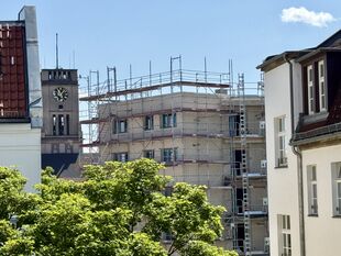 Blick auf ein mit Gerüst umgebenes Gebäude zwischen anderen Häusern und einem Kirchturm mit Uhr im Hintergrund
