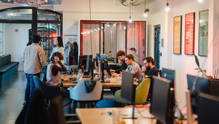 Büro mit mehreren Personen, die an Computern arbeiten, und zwei Personen im Gespräch im Hintergrund