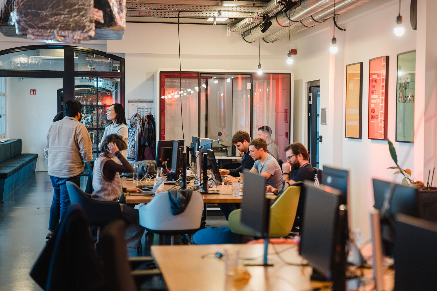 Büro mit mehreren Personen, die an Computern arbeiten, und zwei Personen im Gespräch im Hintergrund