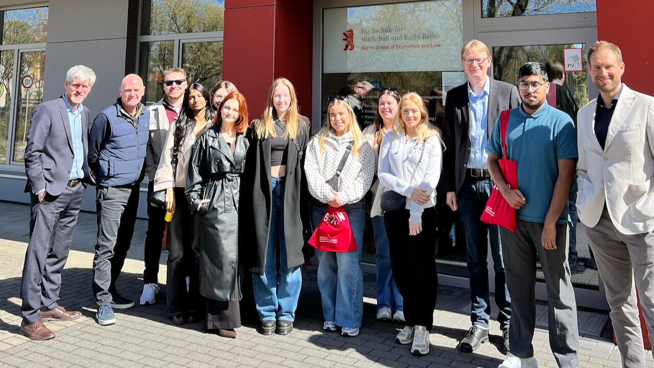Gruppe von Menschen vor einem Gebäude mit Glasfenstern und roter Fassade, einige halten rote Taschen mit weißem Aufdruck