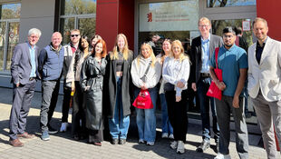 Gruppe von Menschen vor einem Gebäude mit Glasfenstern und roter Fassade, einige halten rote Taschen mit weißem Aufdruck