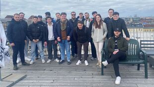 Gruppe von jungen Erwachsenen steht auf einer Holzterrasse mit Stadtblick unter bewölktem Himmel, eine Person sitzt auf einer Bank