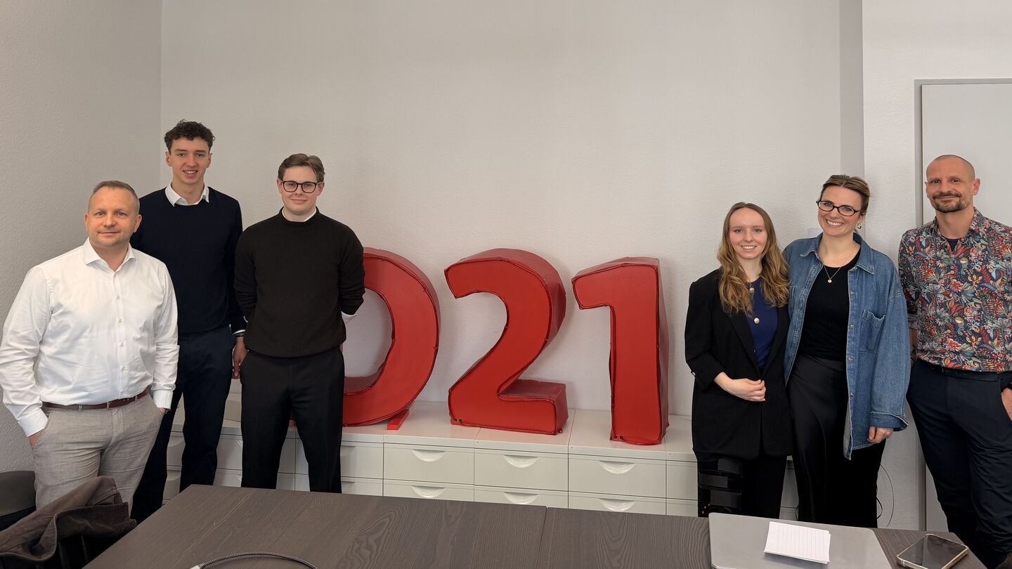 Gruppe von Menschen in einem Besprechungsraum mit einem Bildschirm im Hintergrund und großen roten Zahlen '021' auf einem Sideboard